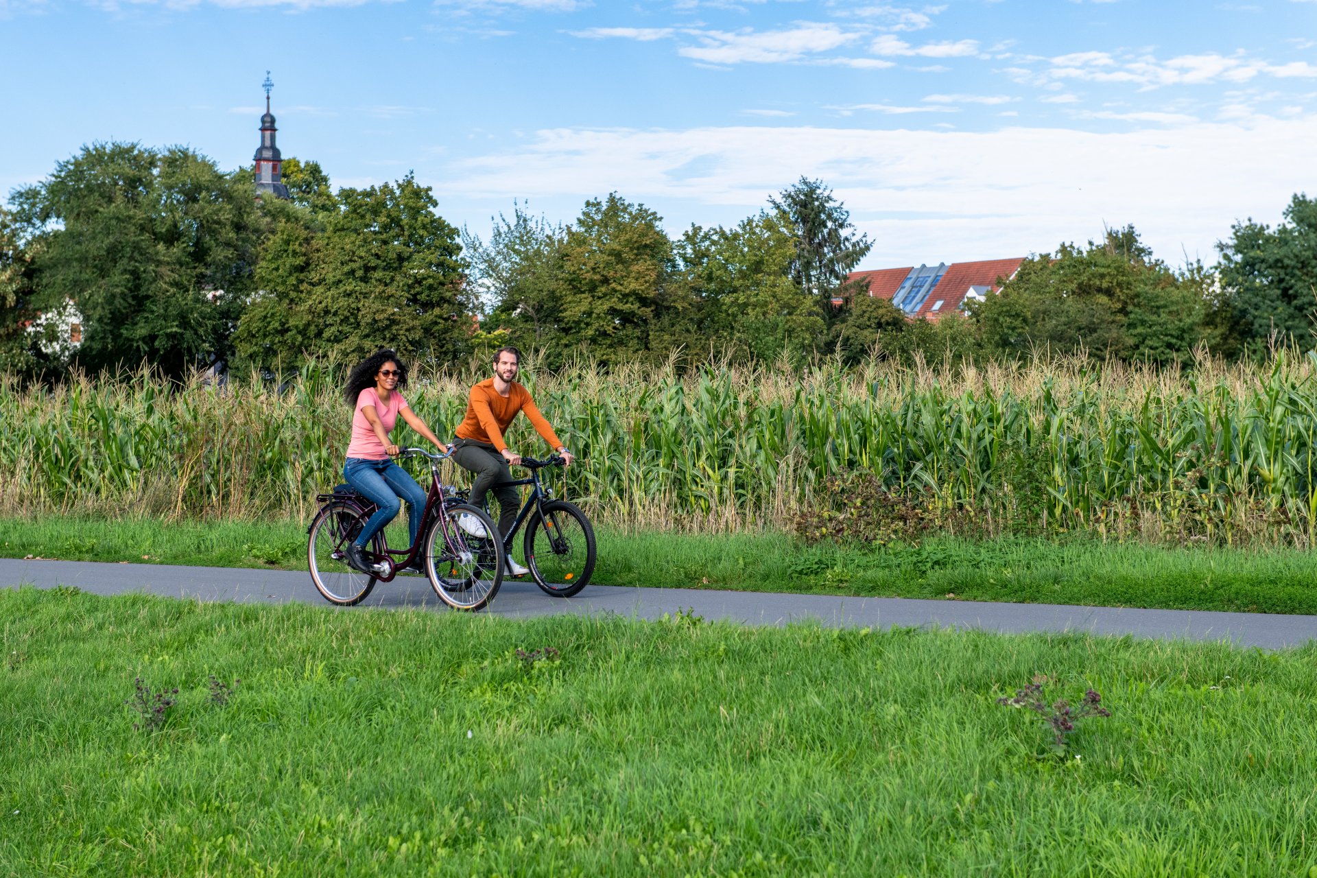 Zwei Personen fahren mit Fahrrädern auf einem Weg, im Hintergrund ist ein Maisfeld.