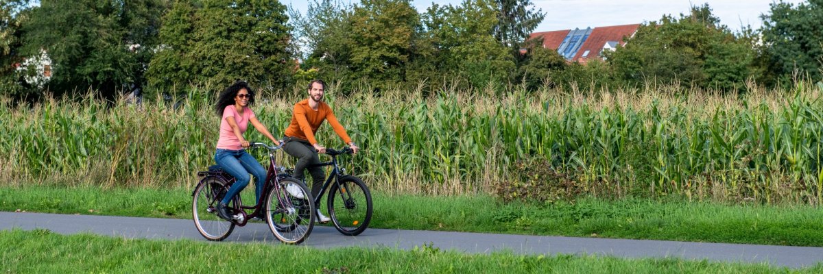 Zwei Personen fahren mit Fahrrädern auf einem Weg, im Hintergrund ist ein Maisfeld.