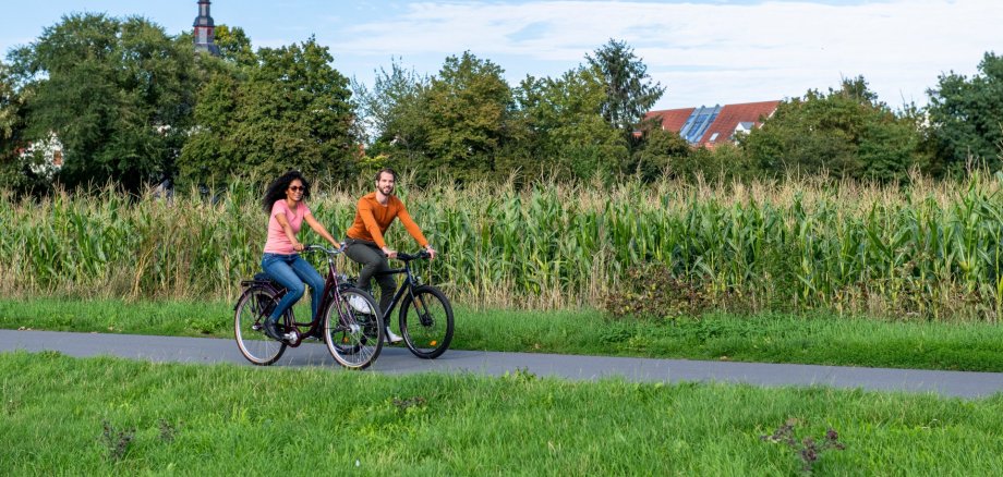 Zwei Personen fahren mit Fahrrädern auf einem Weg, im Hintergrund ist ein Maisfeld.